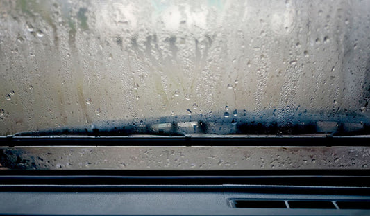 A car windshield covered in raindrops, showcasing a fogged surface contrasted with clear, anti-fog windows.